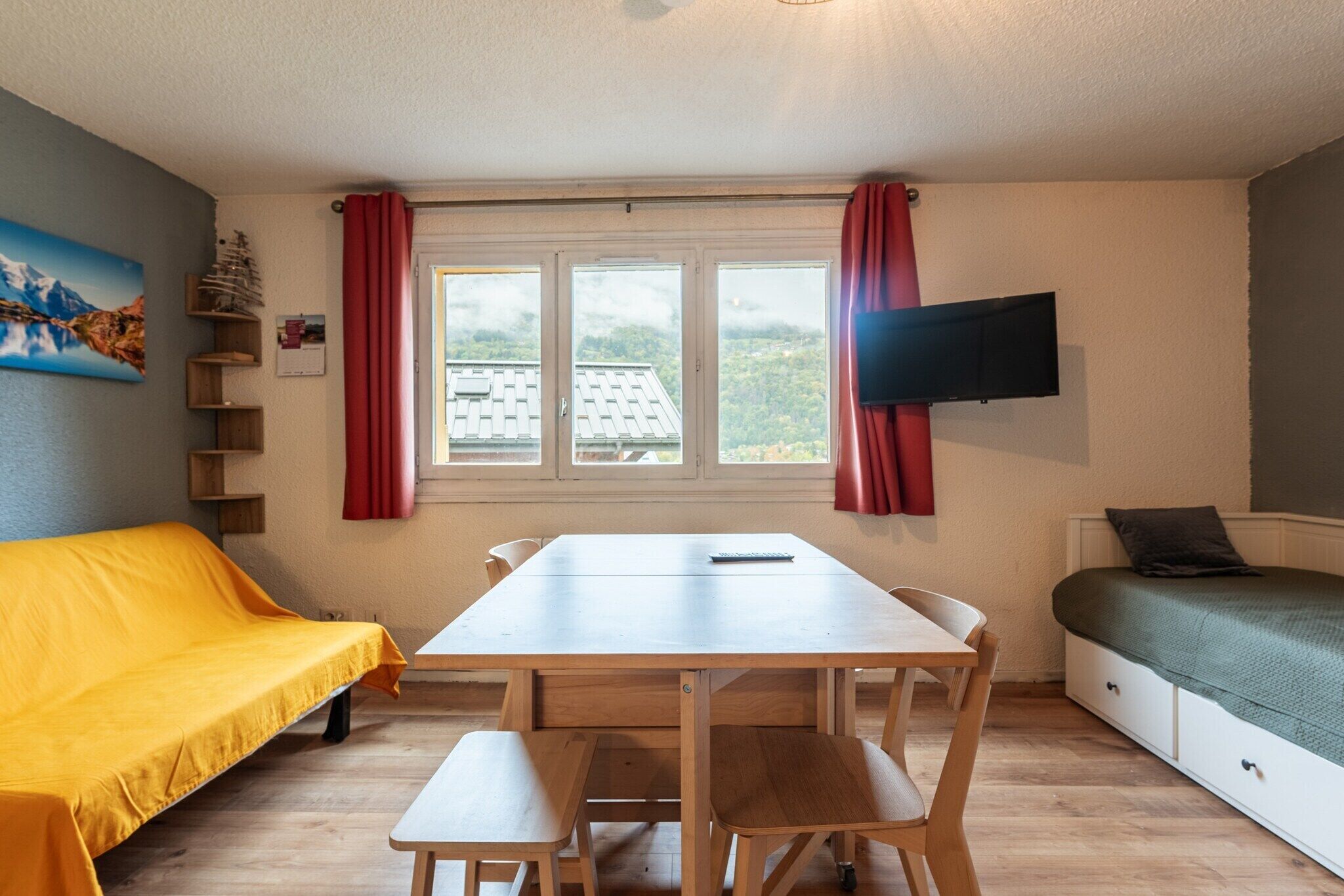 A room with a wooden table, benches, a yellow sofa, a bed, a TV, and a window with curtains.