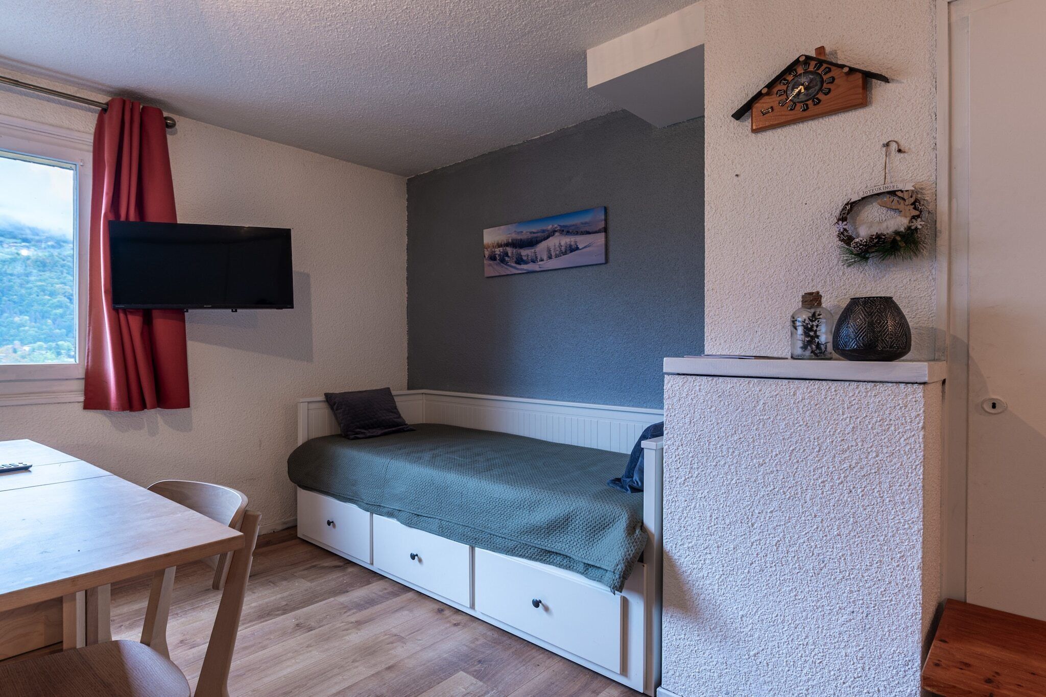 Ein kleines Zimmer mit einem Bett, einem an der Wand befestigten Fernseher, einem kleinen Tisch und einem Fenster mit Vorhängen.