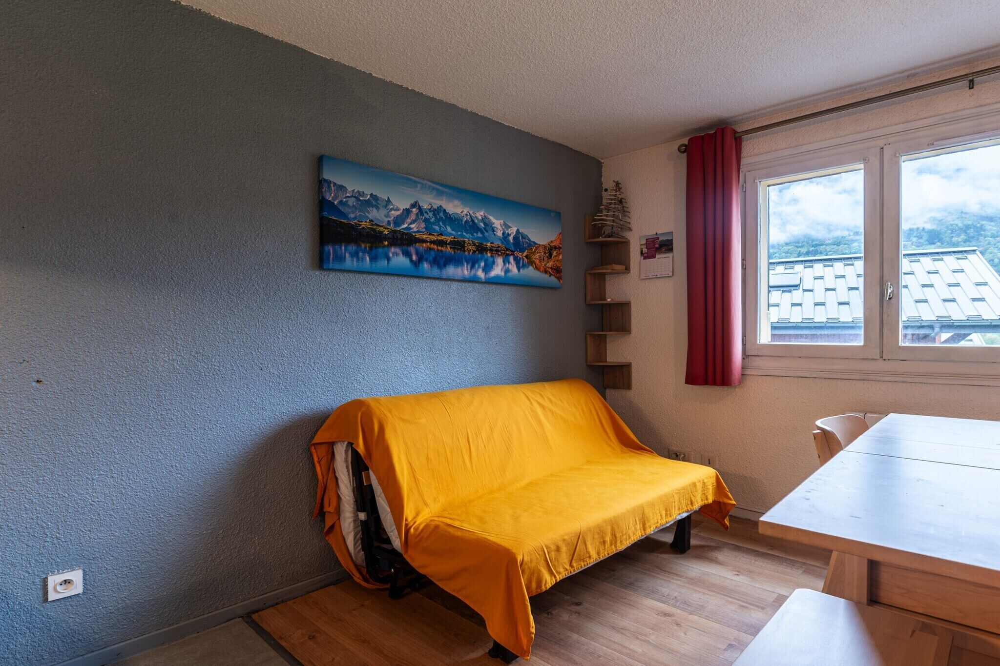 A room with a sofa covered in yellow, a painting of a mountain lake, a window with a red curtain, and a wooden table.