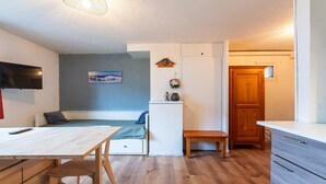 A compact living space with a wooden table, a bench, a sofa, a television, a refrigerator, and a door.