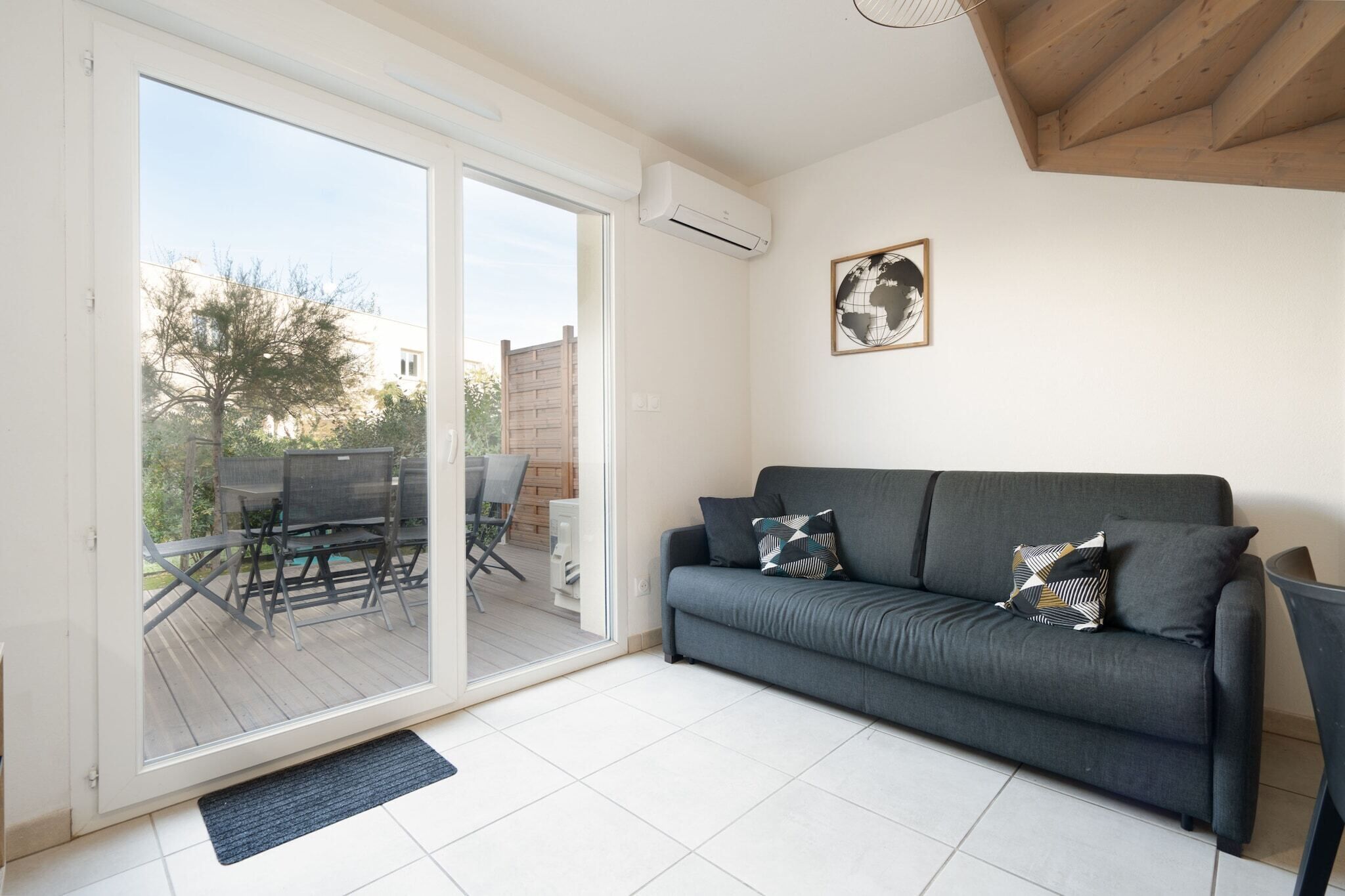 Una sala de estar con un sofá gris, dos cojines estampados, techo de madera y una puerta corrediza de vidrio que da a un patio exterior con mesa y sillas.