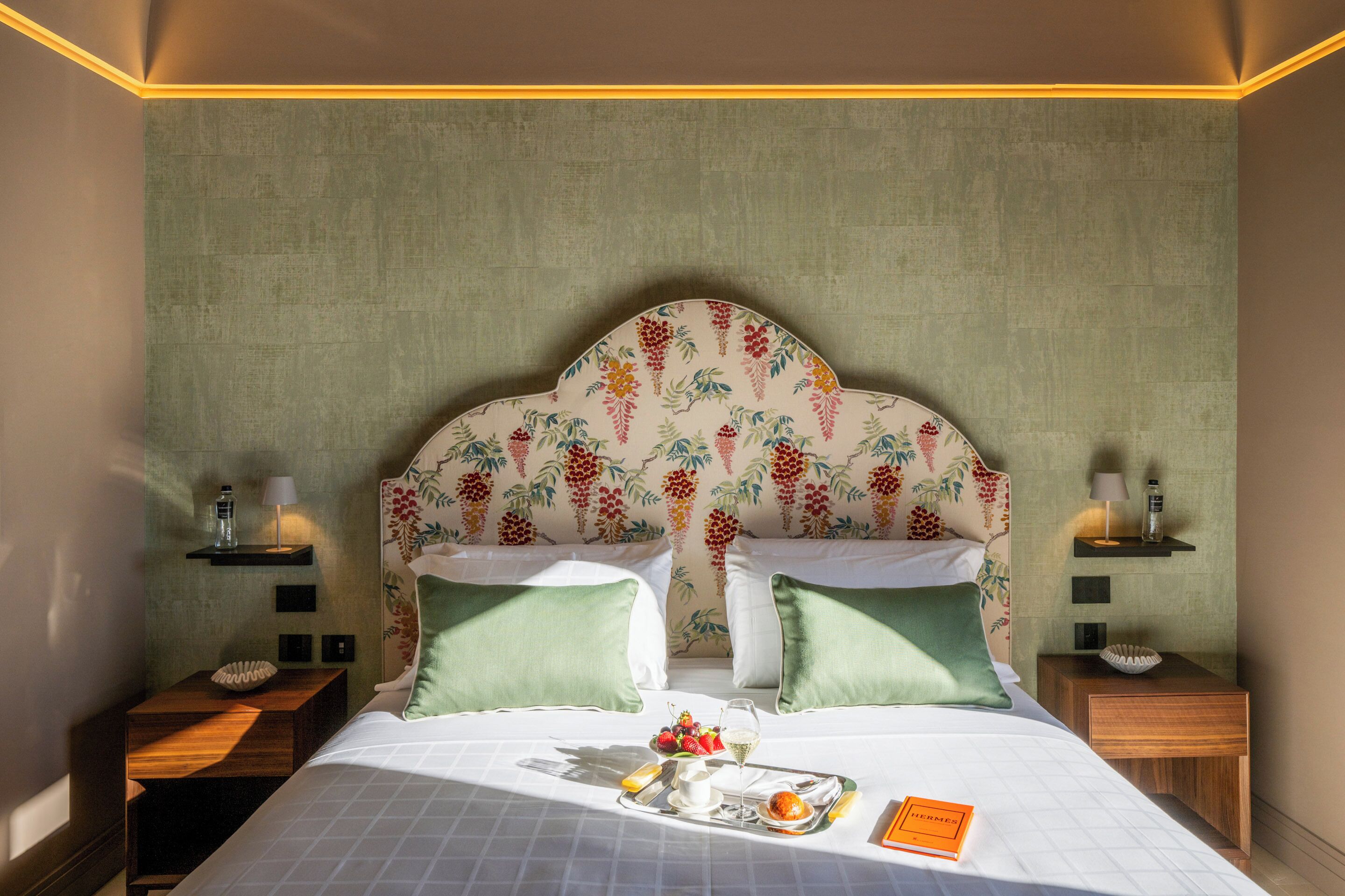 Une chambre d’hôtel avec un lit, des oreillers verts, un plateau de fruits et un verre de vin, ainsi que deux tables de chevet avec des lampes.