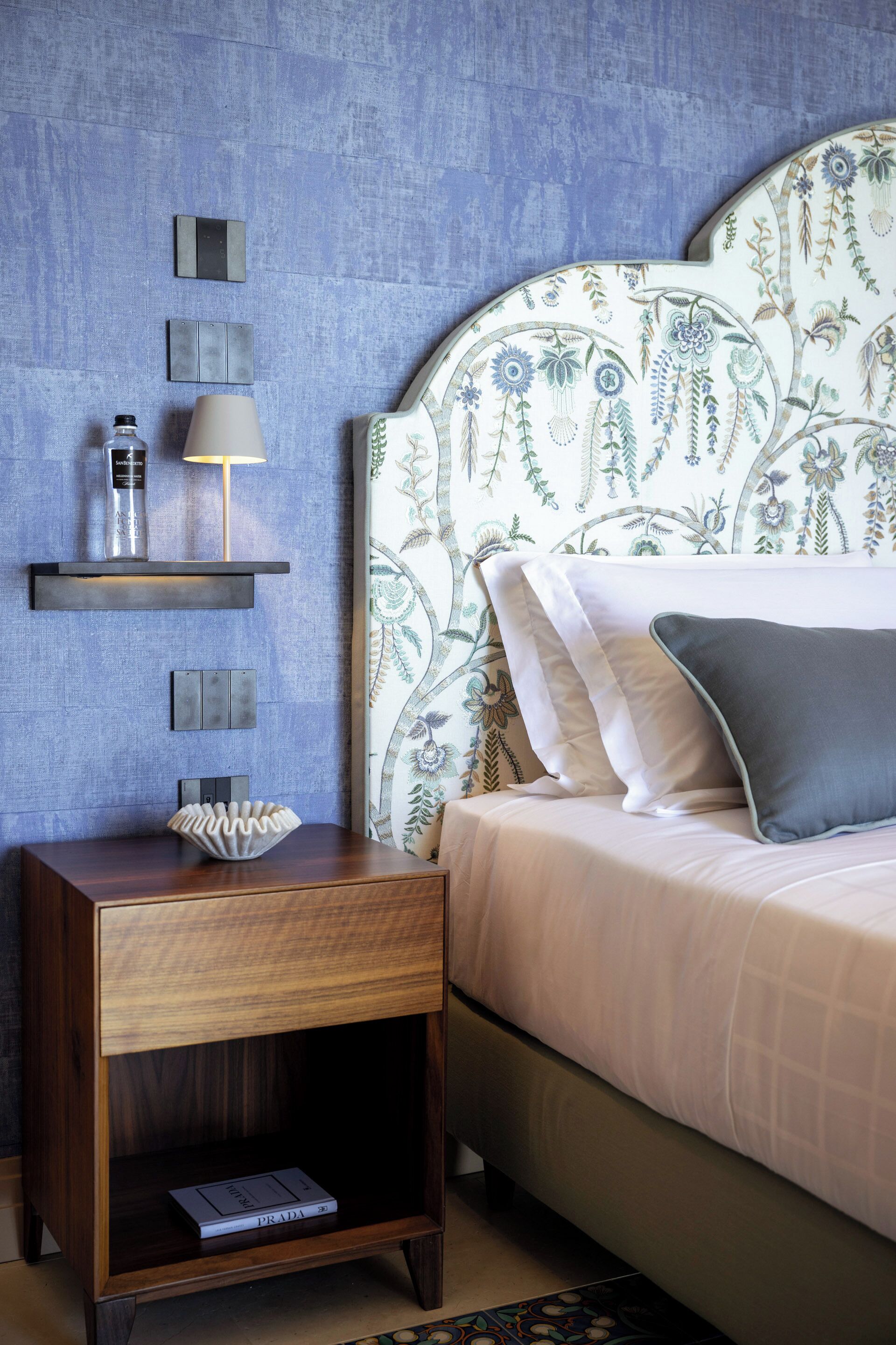 A bedroom with a floral-patterned headboard, a bedside table with a lamp, and a shelf with toiletries.