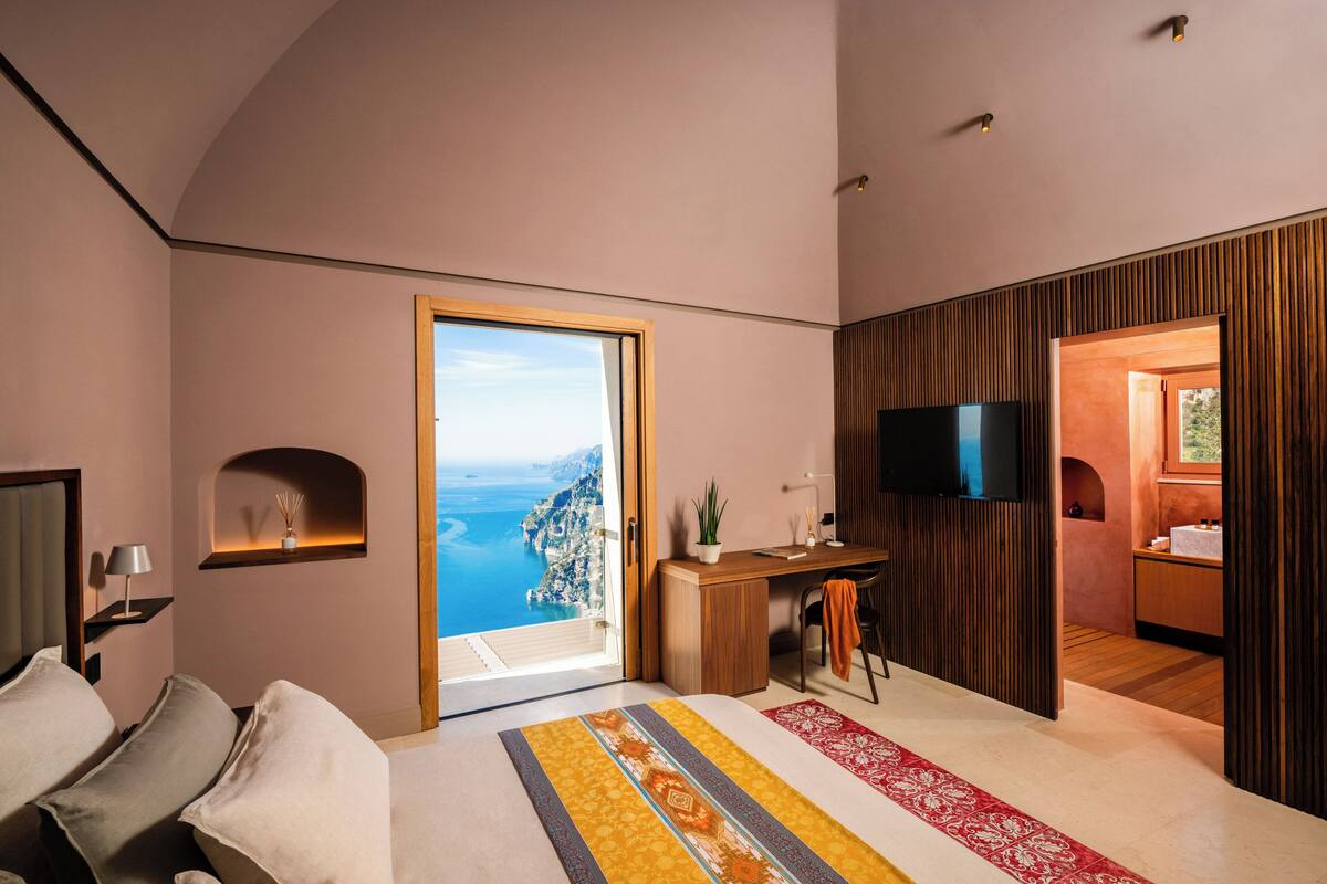Un dormitorio moderno con una cama grande, un escritorio y un televisor. Se ve el océano a través de un ventanal amplio.