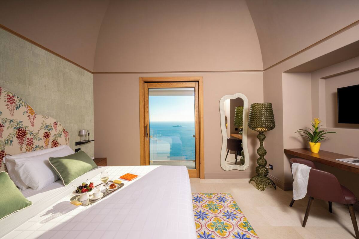 Un dormitorio con cama, escritorio, silla, espejo, planta en maceta y vista al mar.