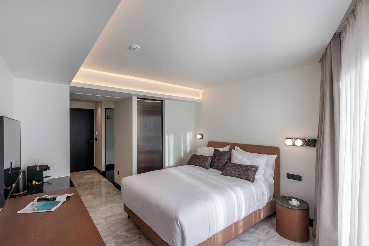 A modern hotel room with a large bed, a desk with a chair, a small round table, and a mirror.