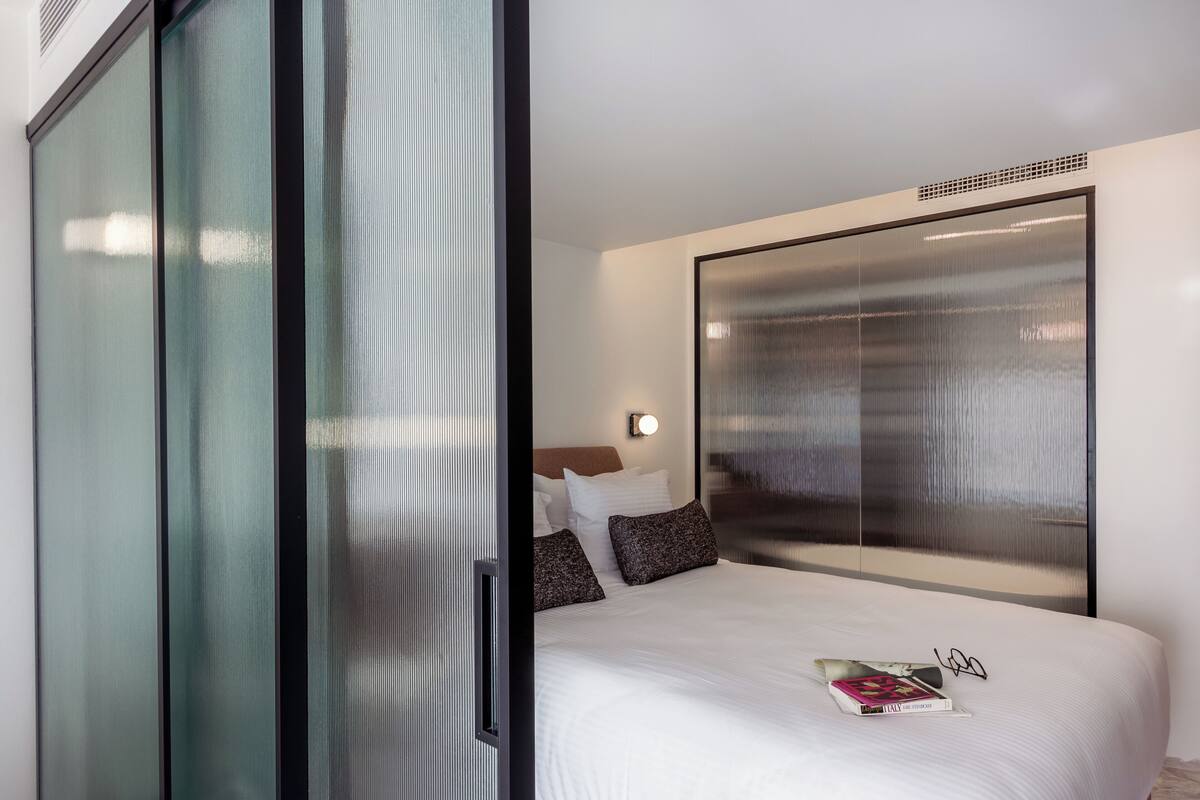 A modern hotel room with a large bed, a glass partition, and a large window.