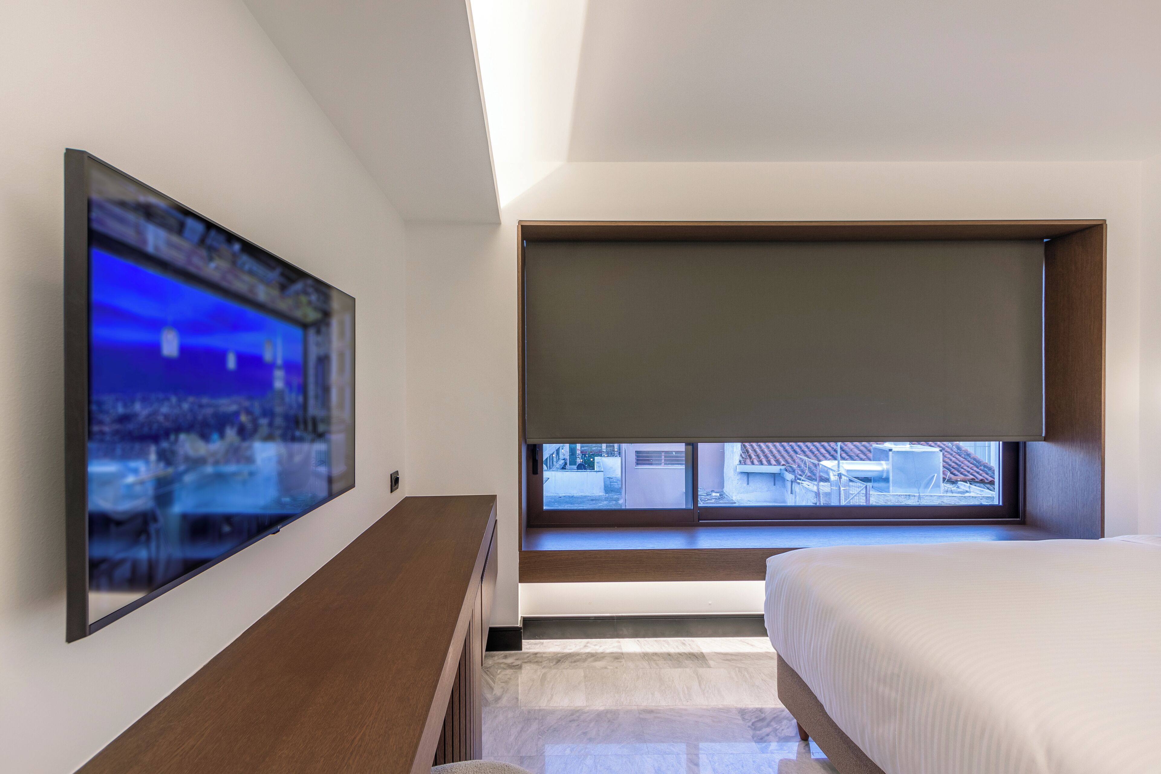 Een moderne slaapkamer met een bed, een aan de muur bevestigde televisie, een raam met uitzicht en een bankje onder het raam.