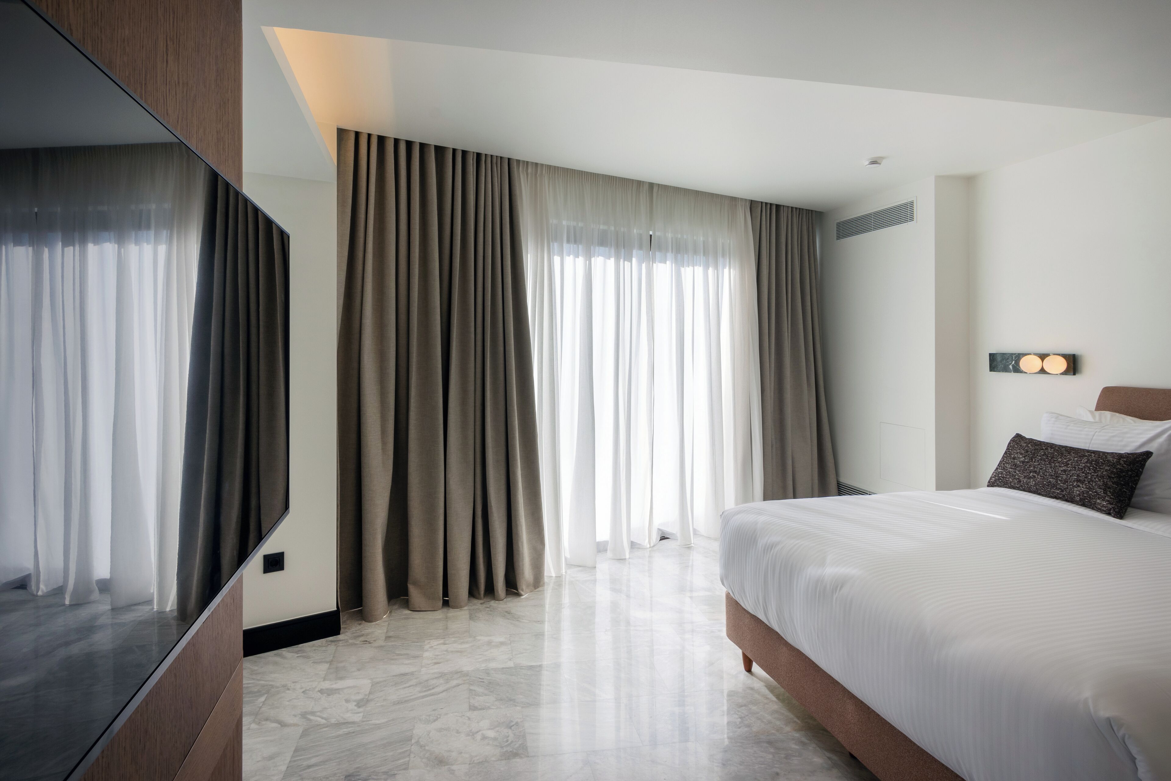 Una habitación de hotel moderna con una cama grande, un televisor de pantalla plana y un amplio ventanal con cortinas.