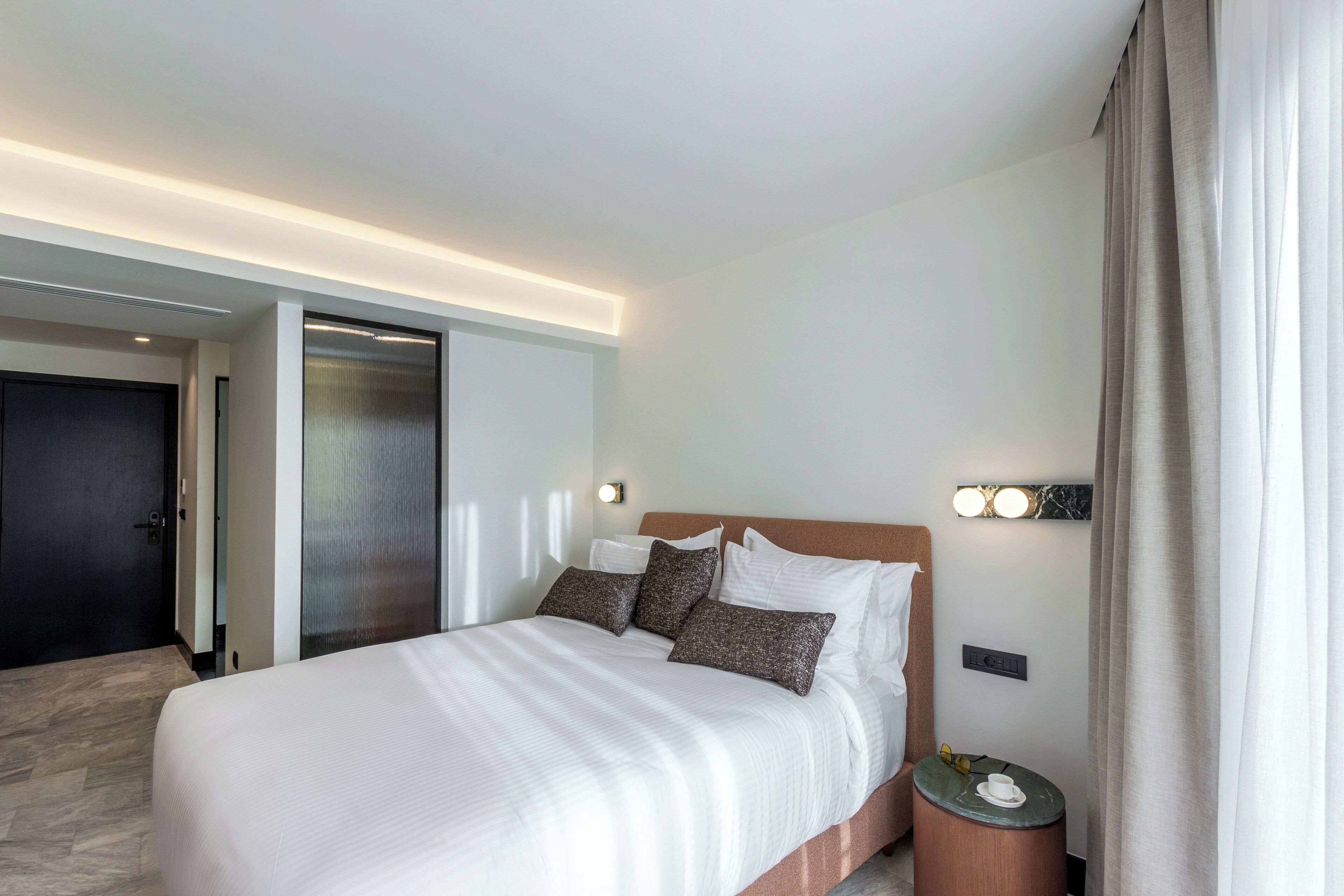 A modern hotel room with a large bed, a bedside table with a lamp, and a view of the hallway.