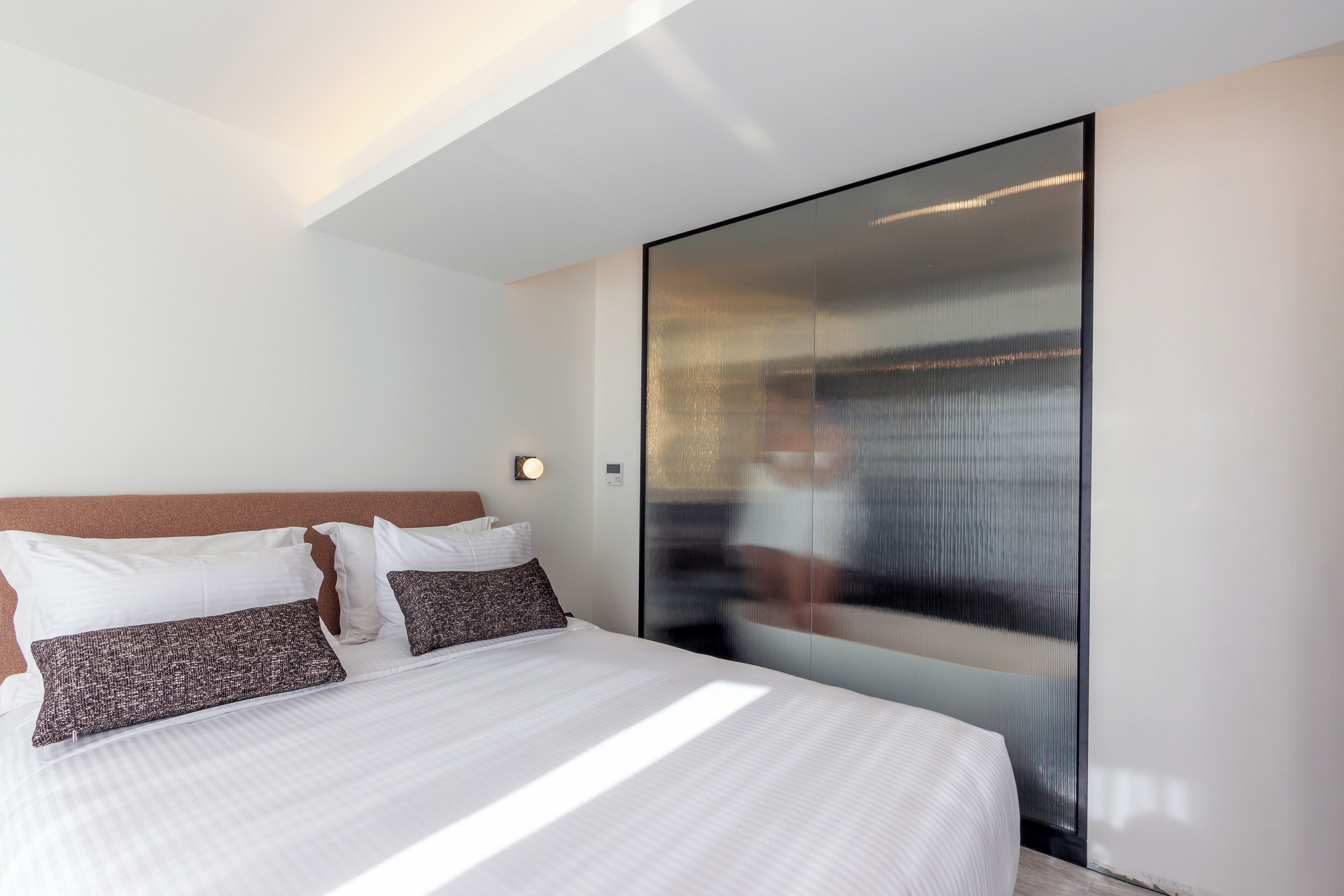 A modern hotel room with a large bed, a glass partition wall, and a ceiling light.