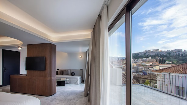 A modern hotel room with a large window, a flat-screen TV, a wooden accent wall, a sofa, and a view of a cityscape.