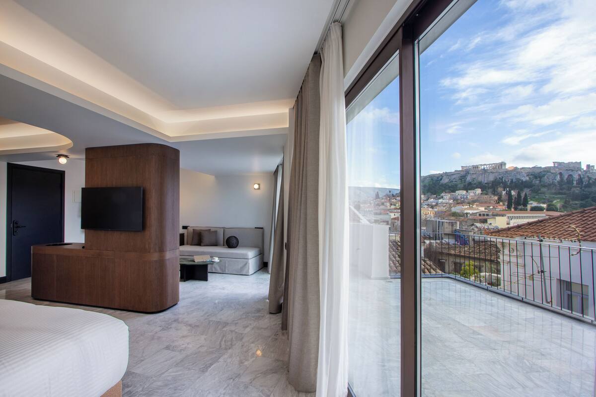 A modern hotel room with a large window, a flat-screen TV, a wooden accent wall, a sofa, and a view of a cityscape.