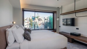 A modern hotel room with a large bed, a TV mounted on the wall, and a view of the cityscape through a large window.