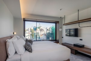 A modern hotel room with a large bed, a TV mounted on the wall, and a view of the cityscape through a large window.