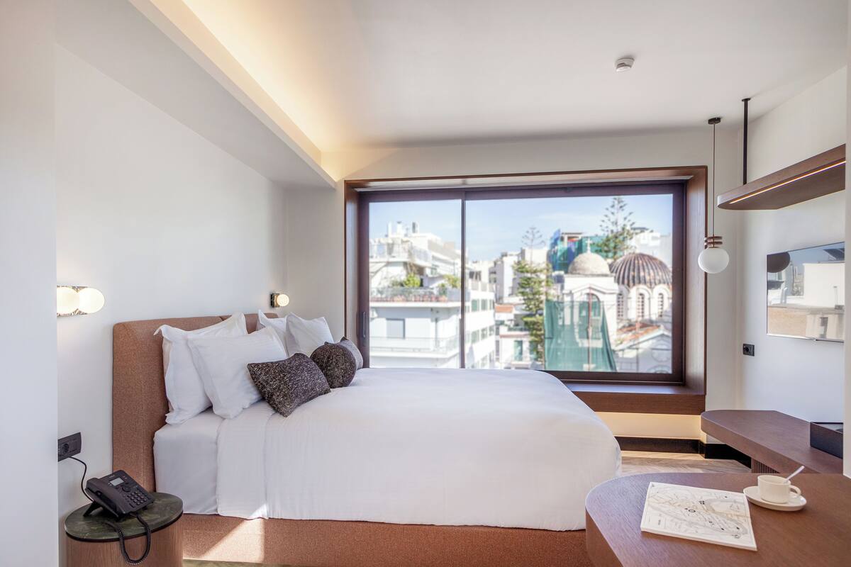 A modern hotel room with a large bed, a bedside telephone, a small table with a cup and saucer, and a view of the city through a large window.