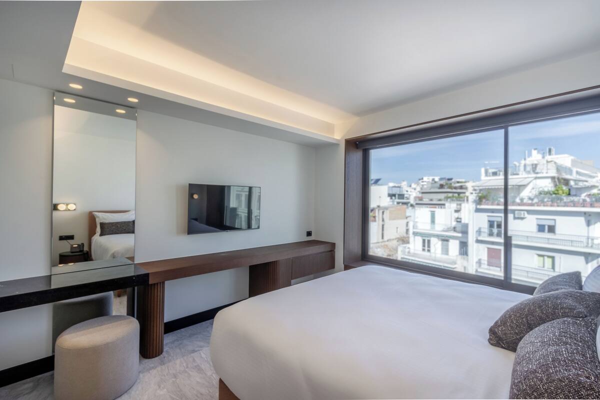 A modern hotel room with a large bed, a TV mounted on the wall, a desk with a chair, and a large window offering a city view.