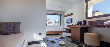 A modern hotel room with a sofa, a coffee table with a book and a cup, a kitchen area with a counter and appliances, and a large window offering a view of the city.