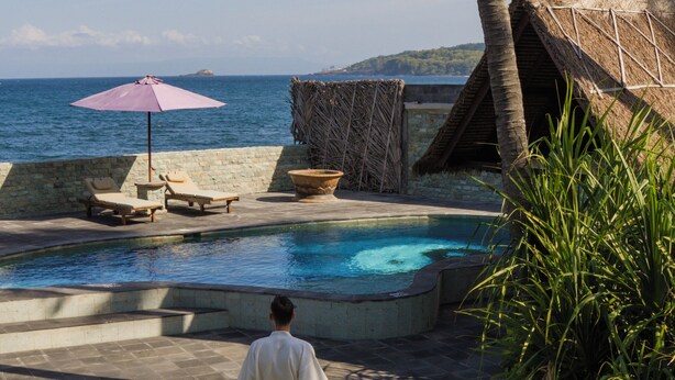 A man walking towards a pool with lounge chairs and a thatched-roof structure. - A hidden gem, away from the hustle and bustle of everyday life. (Karangasem)