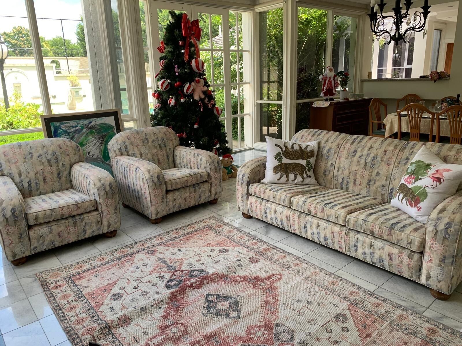 Una sala de estar con un sofá floral, dos sillones, un árbol de Navidad y un comedor con una araña.