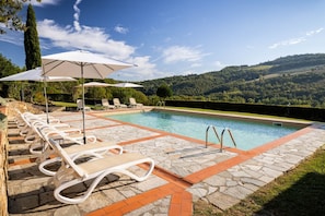 Uma área de piscina com espreguiçadeiras e guarda-sóis, cercada por ladrilhos de pedra e vista para colinas ondulantes.