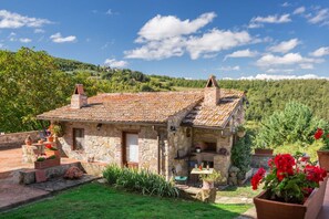 A stone house with a tiled roof, a small garden, and a view of a hilly landscape.