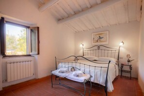 A bedroom with a metal bed frame, white linens, a radiator, a window with curtains, a painting on the wall, and a small table with a lamp. - My Exclusive Italy - Authentic Chianti's Villa with Private Pool and view (Panzano In Chianti)