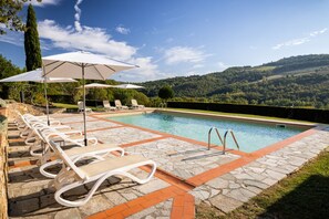 A pool area with lounge chairs and umbrellas, surrounded by stone tiles and a view of rolling hills.