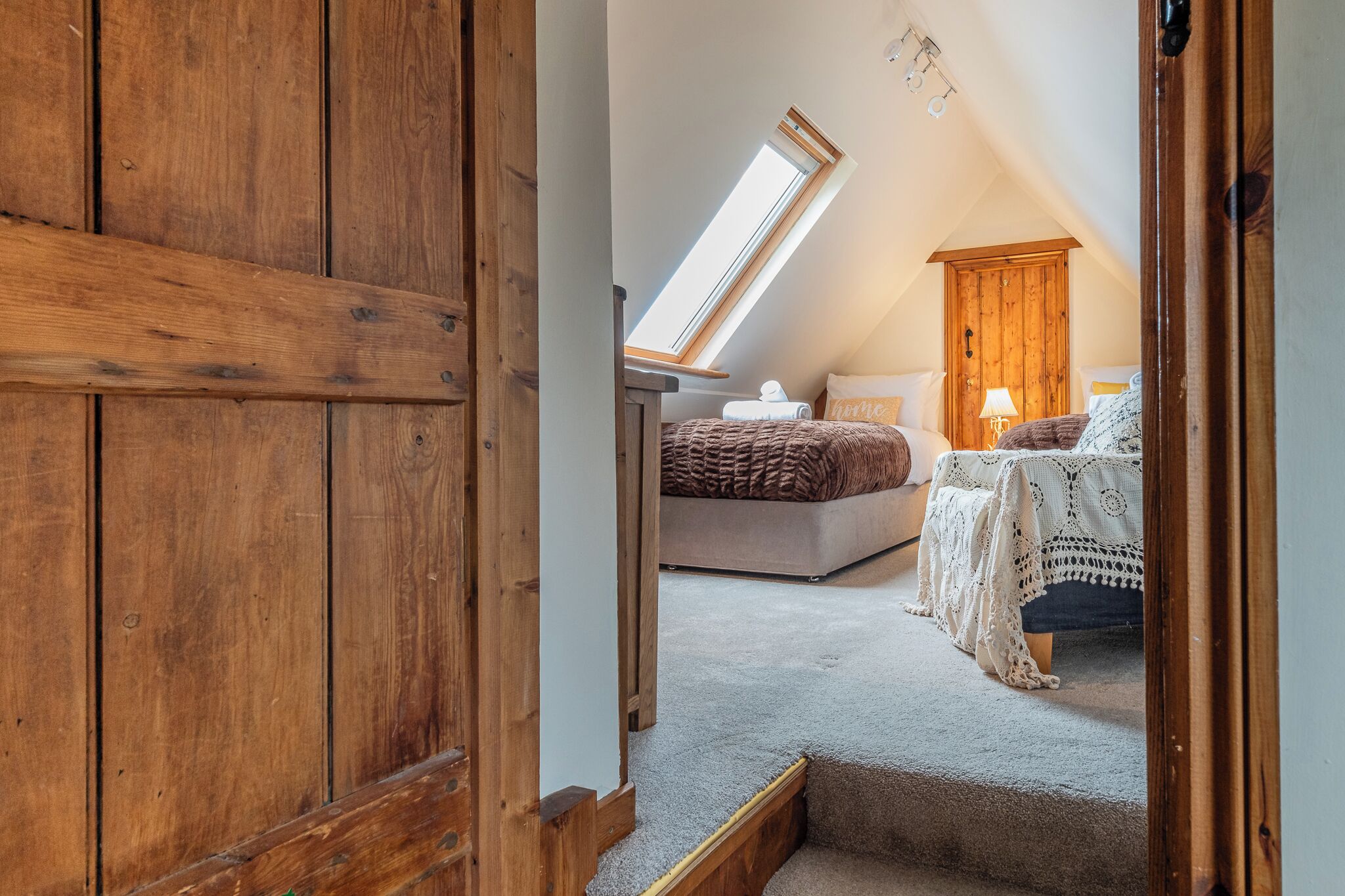 A bedroom with a sloped ceiling, a skylight, a bed with a brown blanket, a bedside table with a lamp, and a wooden wardrobe.