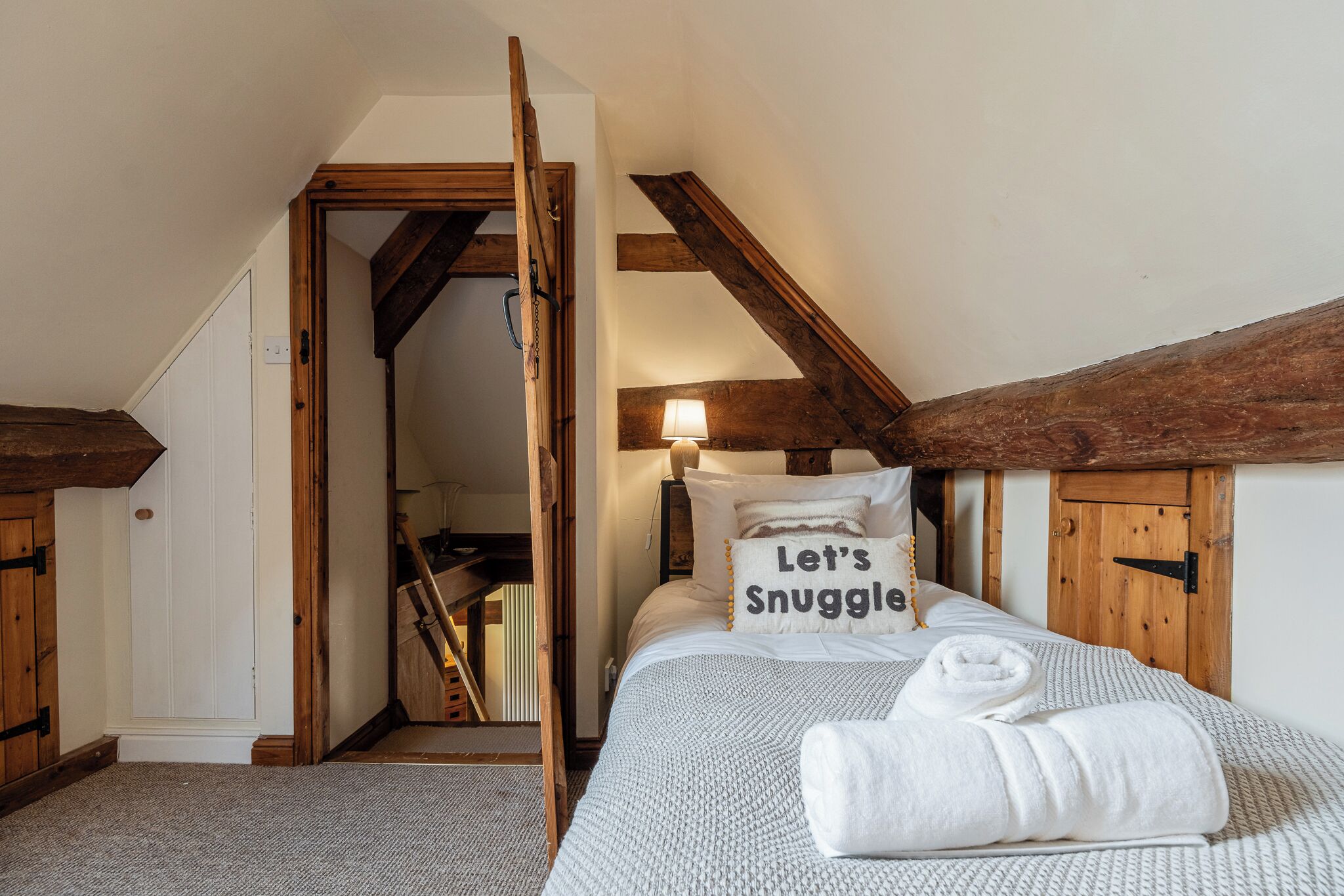 Ein gemütliches Schlafzimmer mit schrägem Dach, Holzbalken, einem Bett mit Kissen mit der Aufschrift 'Let's Snuggle' und einem ordentlich bezogenen Bett mit einem aufgerollten Handtuch.