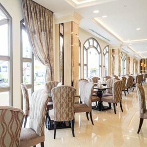 A hotel dining area with multiple tables and chairs, large windows with curtains, and a polished floor. - LK Grand Empire (Pattaya)