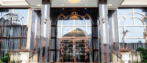 Une grande entrée d’hôtel, avec une vaste porte en arc de cercle, un escalier menant à l’étage supérieur et un luminaire moderne au-dessus.