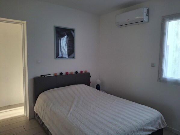 A bedroom with a bed, a wall-mounted air conditioner, a window, and a painting. - Villa Loucantou at Marseillan plage, between Sète and Agde (Marseillan)