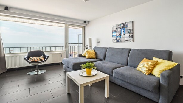 A modern living room with a grey sofa, a small white table with a plant, and a black chair with a patterned cushion.