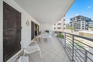 Ein Balkon mit weißen Stühlen und einem Tisch, ein großes Fenster und Blick auf ein modernes Gebäude.