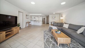 A modern living room with a flat-screen TV, a wooden coffee table, a grey sofa, and a kitchen area in the background.