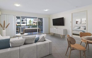 A modern living room with a sofa, a dining table, and a TV mounted on the wall. - Windsong Caloundra: Sunsets Sea Breezes & Style (Caloundra Region)