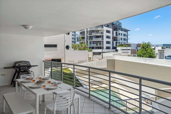 Ein Balkon mit Esstisch für vier Personen, Grill und Blick auf einen Swimmingpool sowie Gebäude.