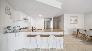 A modern kitchen with white cabinets, a central island with stools, a stainless steel refrigerator, and a dining area with a table and chairs.