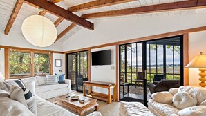 A spacious living room with a large sofa, a coffee table, and a television. There are large windows and a sliding glass door leading to an outdoor area. - Hot Tub & Grill | Folly Marsh House (Folly Beach)