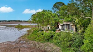 A lakeside cabin with a deck, surrounded by dense greenery and trees. - Hot Tub & Grill | Folly Marsh House (Folly Beach)