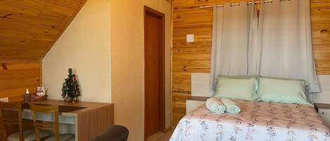 A cozy room with a wooden ceiling, a bed with floral bedding, a small desk with a chair, and a Christmas tree.