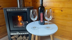 Una estufa de leña con puerta de cristal, una botella de vino y dos copas sobre una mesita blanca.