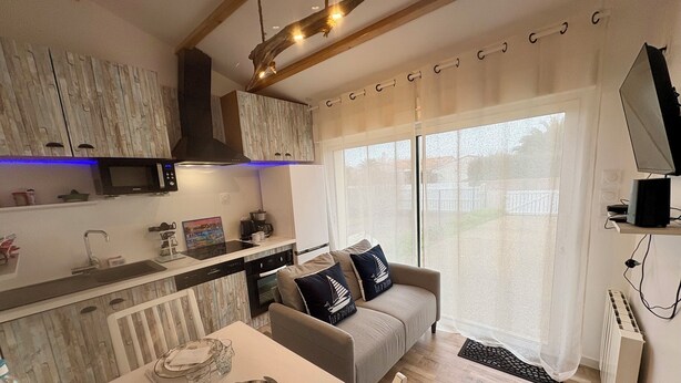 A modern kitchen with a dining area, wooden ceiling beams, and a large window with sheer curtains. - Studio Azur et Mer tout CONFORT (Dolus-d'Oléron)