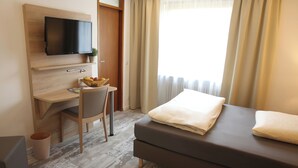 Une chambre d’hôtel moderne dotée d’un téléviseur à écran plat fixé à un mur en bois, d’un bureau avec une chaise, d’une petite plante en pot, d’une table de chevet avec une lampe et d’une grande fenêtre avec des rideaux.