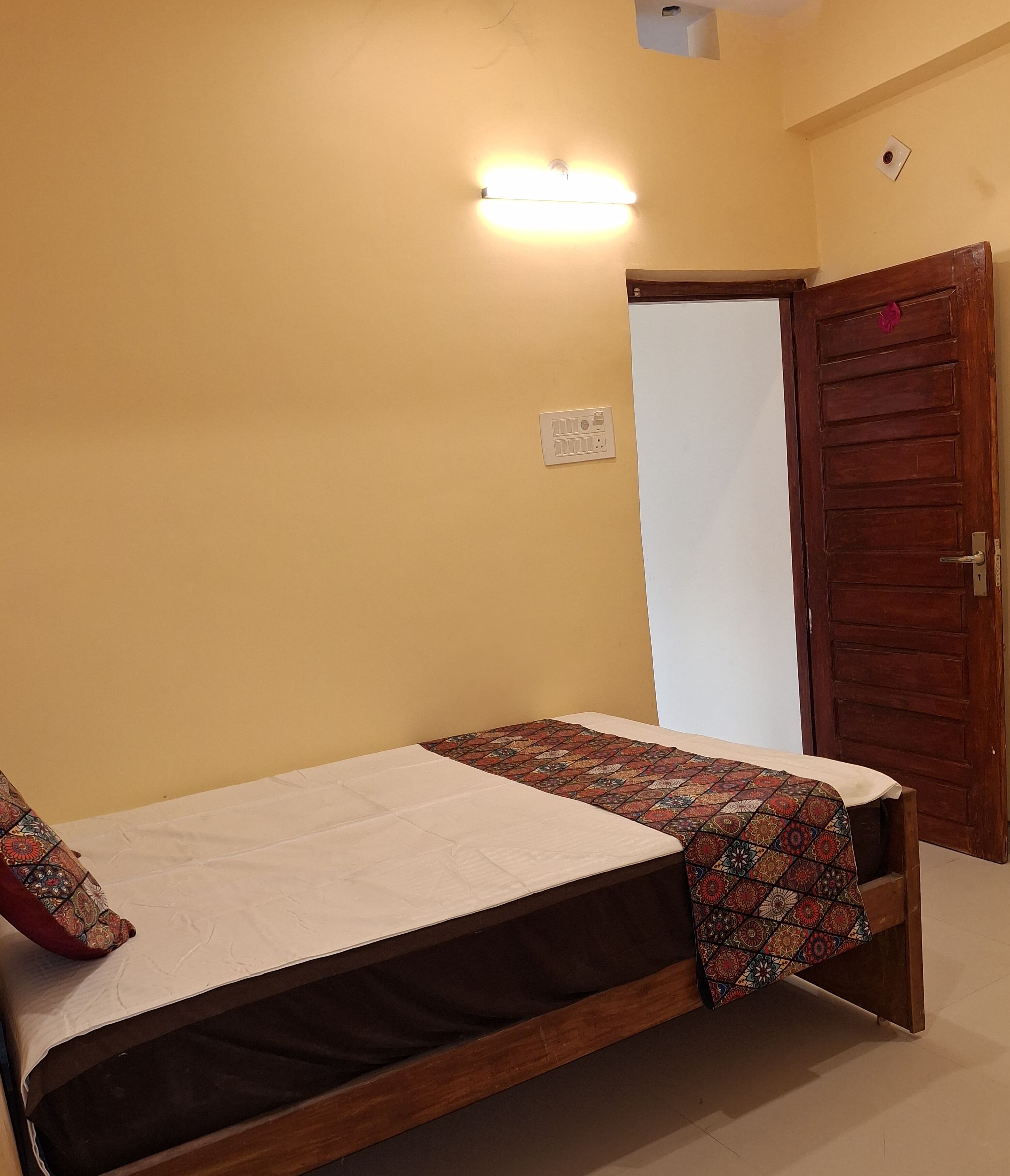 A single bed with a patterned bedspread and pillows, a wooden door, and a wall-mounted light fixture.