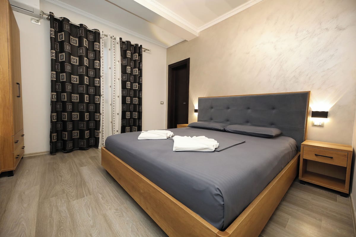 Un lit bien fait, recouvert d’une couvre-lit bleu foncé, avec deux serviettes blanches pliées, une table de chevet en bois dotée d’un tiroir et un luminaire fixé au mur.