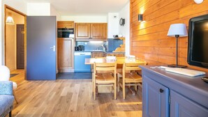 A compact kitchen with wooden cabinets, a dining table with chairs, a blue refrigerator, and a mounted television.