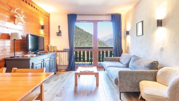 A cozy living room with a wooden wall, a television, a sofa, a dining table, and a balcony with a mountain view.