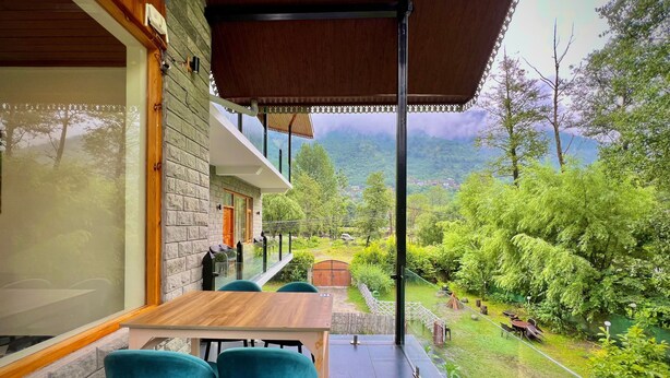 A covered outdoor seating area with a table and chairs, a stone wall, and a view of a lush garden and mountains.